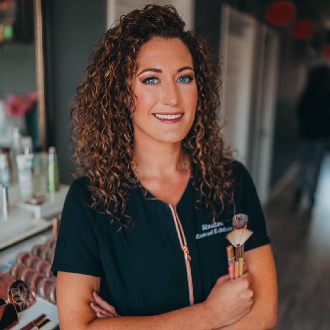 A portrait of a smiling woman with curly brown hair wearing a black short-sleeve zip-front uniform with a small white logo on the chest, standing indoors with her arms crossed. The photo is shown within a mobile app interface with a white surrounding frame and icons.